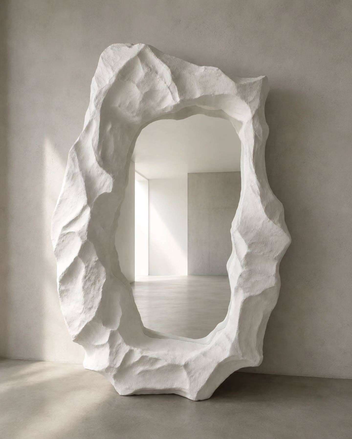 Umbral: espejo escultórico de 1,90 m, terminación mineral blanca y apertura central tallada, apoyado contra un muro de hormigón.
